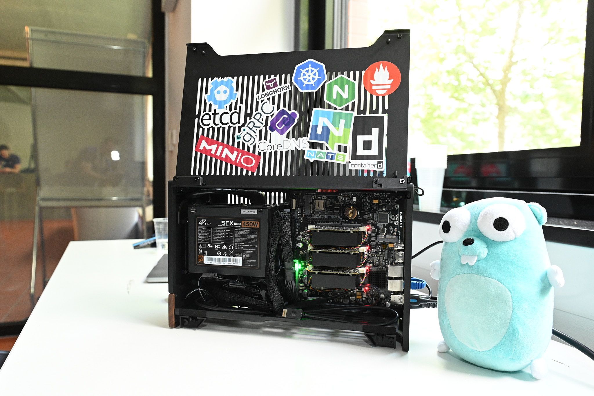 Otevřená počítačová skříň se samolepkami na otevřeném víku a vedle Gopher plyšák (maskot Golang)"